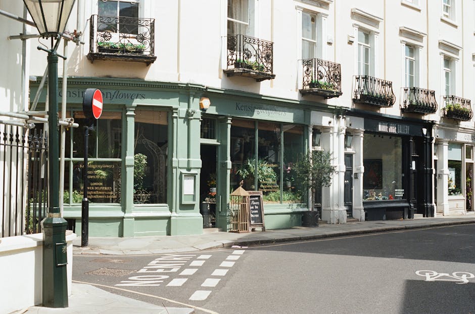 Exterior view of a florist shop on Coldharbour Lane Brixton, with a pastel green storefront featuring large glass windows displaying floral arrangements inside. The shop's signage reads 'Kensington Fl