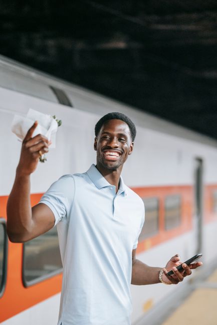 A smiling young man with dark skin and short black hair is holding a small bouquet of flowers wrapped in white paper, standing on a platform at Brixton Station SW9. He is dressed in a light blue polo 