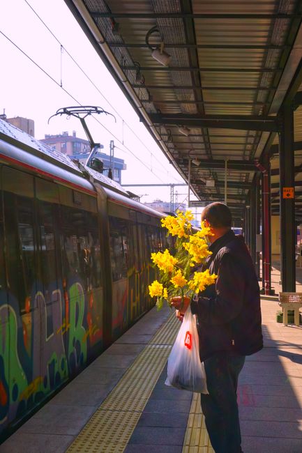 A man standing on Brixton Station SW9 platform holding a bouquet of bright yellow flowers, including daffodils, with fresh, vibrant petals and green stems. He is dressed in a dark jacket and carrying 